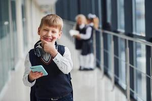 Little boywith phone and headphones standing if front of school kids in uniform that together in corridor. Conception of education photo