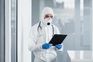 Male doctor scientist in lab coat, defensive eyewear and mask holding notepad in hands photo