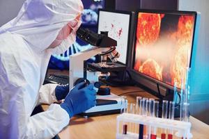 científico con uniforme protector blanco cerca de pantallas con datos trabaja con coronavirus y tubos de sangre en laboratorio foto