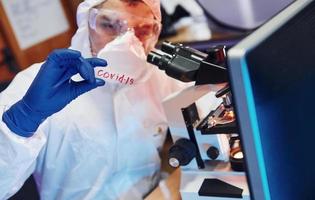 Scientist in white protective wear indoors in laboratory with lens with COVID-19 title on it photo