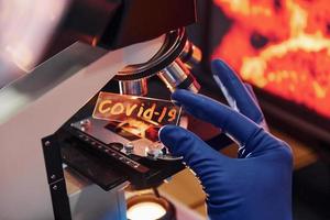 Scientist in white protective wear indoors in laboratory with lens with COVID-19 title on it photo