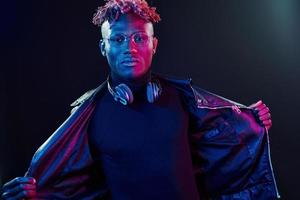 Posing with wireless headphones on neck. Futuristic neon lighting. Young african american man in the studio photo