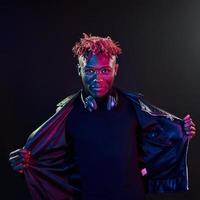 Posing with wireless headphones on neck. Futuristic neon lighting. Young african american man in the studio photo