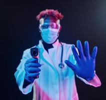 Smart doctor in protective uniform holding infrared thermometer. Futuristic neon lighting. Young african american man in the studio photo