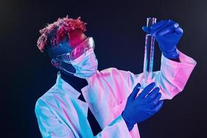 Smart scientist in protective uniform holding test tube. Coronavirus conception. Futuristic neon lighting. Young african american man in the studio photo