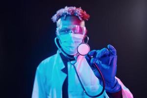 Smart doctor in protective uniform holding stethoscope. Futuristic neon lighting. Young african american man in the studio photo