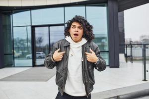 Handsome young man with curly black hair posing for the camera on the street against building photo