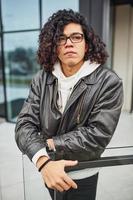 Handsome young man with curly black hair and in glasses is on the street against building photo