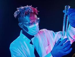 científico inteligente con uniforme protector que sostiene el tubo de ensayo. concepción del coronavirus. iluminación de neón futurista. joven afroamericano en el estudio foto