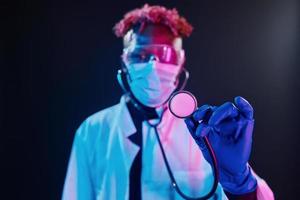 Smart doctor in protective uniform holding stethoscope. Futuristic neon lighting. Young african american man in the studio photo