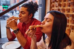 pareja joven multiétnica sentada en el interior juntos y comiendo pizza foto