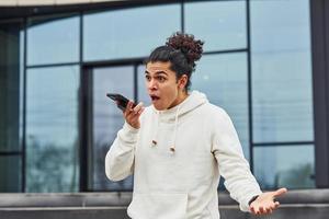 un joven guapo con el pelo negro y rizado está en la calle contra el edificio hablando por teléfono foto