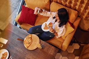Young brunette sitting indoors with pizza and smartphone in hand photo