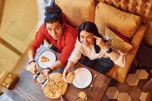 vista superior de una pareja joven multiétnica que se sientan juntos en el interior y comen pizza foto