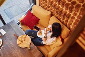 Young brunette sitting indoors with pizza and smartphone in hand photo