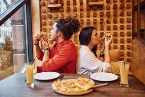 Multi ethnic young couple sitting indoors together and eating pizza photo