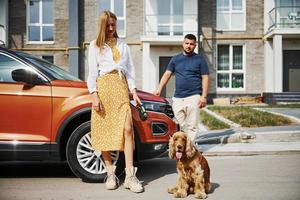 Going out from the building. Lovely couple have a walk together with dog outdoors near the car photo