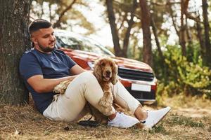 el hombre se sienta cerca del árbol con su perro al aire libre en el bosque foto