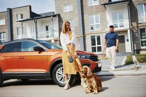 Going out from the building. Lovely couple have a walk together with dog outdoors near the car photo