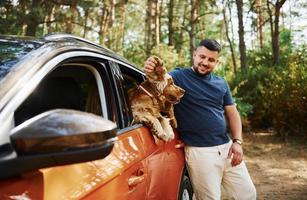 Man stands by the car in the forest. Cute dog in vehicle looks through the window photo