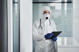 Male doctor scientist in lab coat, defensive eyewear and mask holding notepad in hands photo