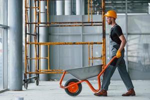 Walks with wheelbarrows. Man in grey uniform works indoors in modern big office at daytime photo