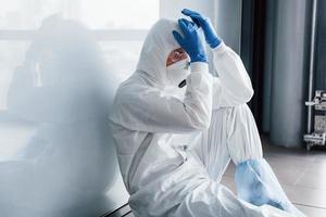 Tired doctor scientist in lab coat, defensive eyewear and mask sits and takes break photo