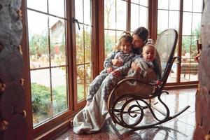 Happy mother sits with her little son and daughter on the chair near window photo
