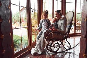 Happy mother sits with her little son and daughter on the chair near window photo