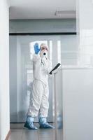 Shows stop gesture by hand. Male doctor scientist in lab coat, defensive eyewear and mask holding notepad in hands photo