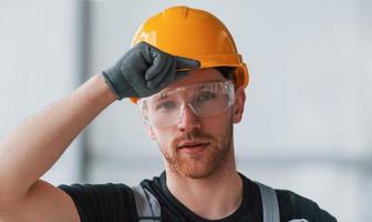 Portrait of man in grey uniform, protective glasses and orange hard hat that standing indoors in modern big office at daytime photo