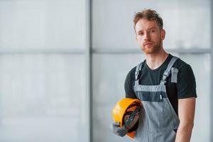 retrato de un hombre con uniforme gris y un casco naranja en las manos que está parado en el interior de una gran oficina moderna durante el día foto