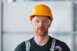 retrato de un hombre con uniforme gris, gafas protectoras y un casco naranja que se encuentra en el interior de una gran oficina moderna durante el día foto