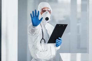 Shows stop gesture by hand. Male doctor scientist in lab coat, defensive eyewear and mask holding notepad in hands photo