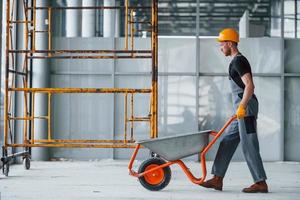 Walks with wheelbarrows. Man in grey uniform works indoors in modern big office at daytime photo