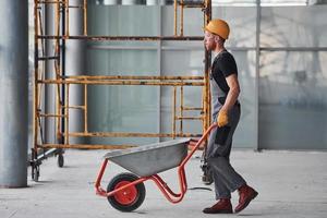 Walks with wheelbarrows. Man in grey uniform works indoors in modern big office at daytime photo