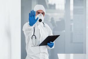 Shows stop gesture by hand. Male doctor scientist in lab coat, defensive eyewear and mask holding notepad in hands photo