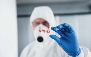 Male doctor scientist in lab coat, defensive eyewear and mask holds glass with virus word on it photo