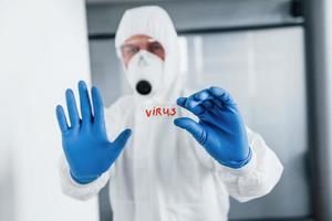 Male doctor scientist in lab coat, defensive eyewear and mask holds glass with virus word on it photo