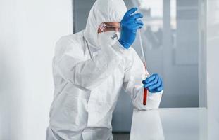 Male doctor scientist in lab coat, defensive eyewear and mask works with test tube with blood inside of it photo