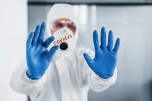 Male doctor scientist in lab coat, defensive eyewear and mask holds glass with covid-19 word on it photo