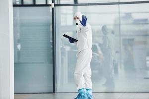 Shows stop gesture by hand. Male doctor scientist in lab coat, defensive eyewear and mask holding notepad in hands photo