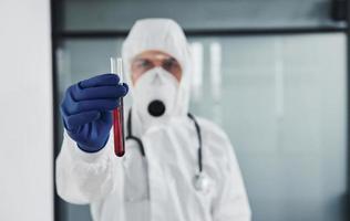 médico científico masculino en bata de laboratorio, anteojos defensivos y máscara sostiene tubo de ensayo con sangre foto
