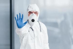 Shows stop gesture by hand. Male doctor scientist in lab coat, defensive eyewear and mask photo