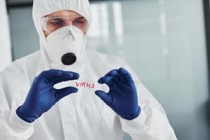 médico científico masculino en bata de laboratorio, anteojos defensivos y máscara sostiene vidrio con palabra de virus en él foto