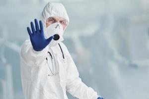 Shows stop gesture by hand. Male doctor scientist in lab coat, defensive eyewear and mask photo