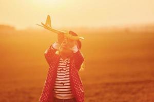 iluminado por la luz del sol de color naranja. una niña linda se divierte con un avión de juguete en el hermoso campo durante el día foto