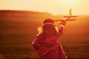 Illuminated by orange colored sunlight. Cute little girl have fun with toy plane on the beautiful field at daytime photo