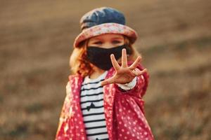 Showing hand. Portrait of cute little girl in black protective mask that standing outdoors photo