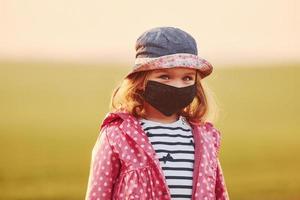 Portrait of cute little girl in black protective mask that standing outdoors illuminated by orange sunlight photo